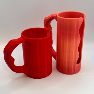 May include: Two red mugs with unique handle designs. One mug is a solid red, while the other has a gradient from red to orange. Both mugs have vertical ridges and are set against a white background.