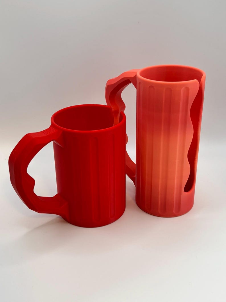 May include: Two red mugs with unique handle designs. One mug is a solid red, while the other has a gradient from red to orange. Both mugs have vertical ridges and are set against a white background.