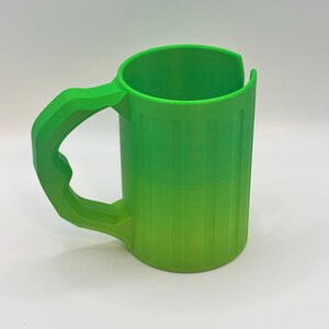 May include: A bright green mug with a large handle. The mug has vertical lines on the side and a curved top. The handle is thick and angular, matching the mug's vibrant color. The mug is made of plastic.