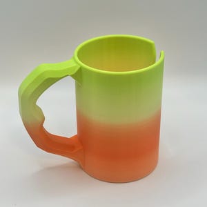 May include: A 3D-printed mug with a gradient design. The mug transitions from a bright neon green at the top to a vibrant orange at the bottom. The handle is a textured, angular design, matching the green color.