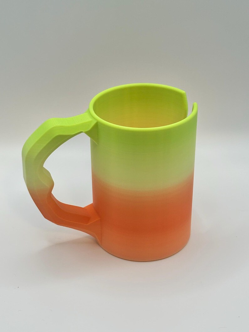 May include: A 3D-printed mug with a gradient design. The mug transitions from a bright neon green at the top to a vibrant orange at the bottom. The handle is a textured, angular design, matching the green color.
