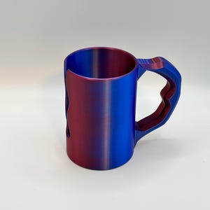 May include: A two-tone mug with a unique handle design. The mug transitions from a deep red to a vibrant blue, creating a gradient effect. The handle has a geometric shape, adding a modern touch to the design. The mug is made of a solid material.