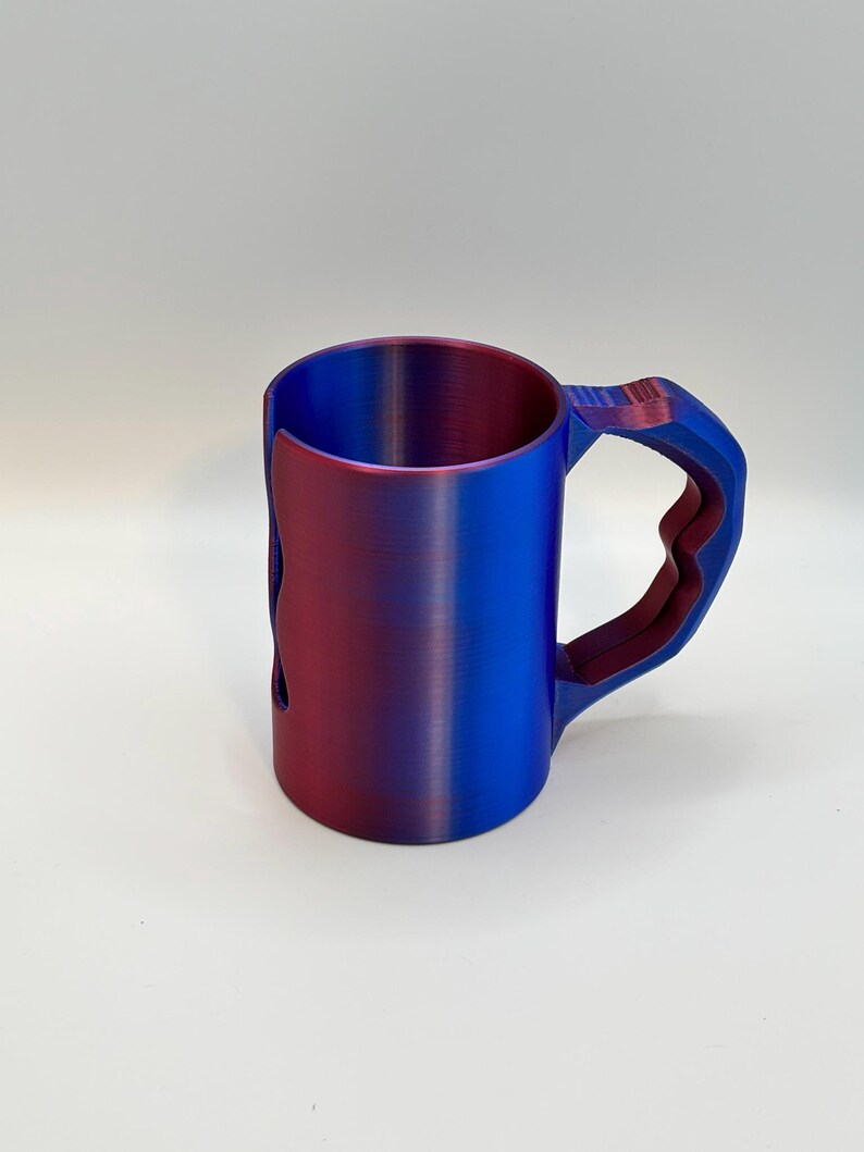May include: A two-tone mug with a unique handle design. The mug transitions from a deep red to a vibrant blue, creating a gradient effect. The handle has a geometric shape, adding a modern touch to the design. The mug is made of a solid material.