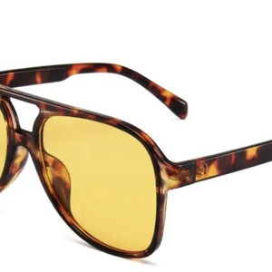 May include: Aviator-style sunglasses with a tortoiseshell frame and yellow-tinted lenses. The sunglasses have a double bridge and thick temples. The lenses are a bright yellow color, and the frame has a brown and black pattern.