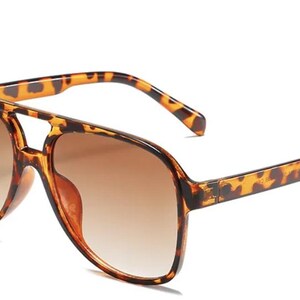May include: Brown gradient lens sunglasses with a tortoise shell pattern on the frames and arms. The sunglasses have a double bridge design and a classic aviator shape. The lenses transition from a darker brown at the top to a lighter brown at the bottom.