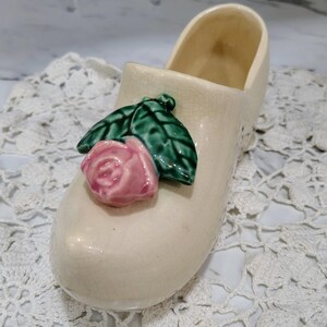 May include: A cream-colored ceramic clog figurine featuring a glossy finish with a fine crackle pattern. The top of the shoe is decorated with a three-dimensional sculpted pink rose blossom surrounded by two dark green leaves with visible veins.
