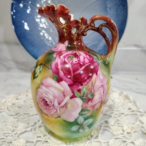 May include: A porcelain pitcher featuring a ruffled rim with gold accents and a curved handle. The body is decorated with large pink and deep red rose blossoms against a yellow and green background, with small cutouts near the top rim.