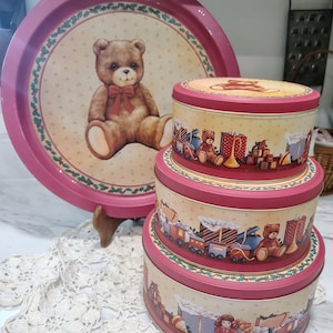 May include: A set of three round metal nesting tins and a matching round metal tray. The tins and tray feature a cream background with a red border, decorated with illustrations of teddy bears, toys, gift boxes, and green holly leaf accents.