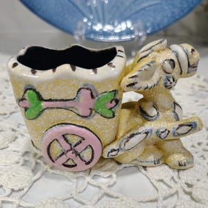 May include: A yellow ceramic donkey figurine pulling a cart with a pink wheel and a green and pink arrow design. The donkey has white and black accents, and the cart features a scalloped top edge with a dark interior, finished in a speckled glaze.