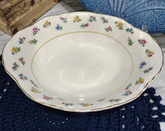 May include: An oval, white porcelain serving bowl with a gold rim and floral pattern. The rim is scalloped and decorated with small, colorful flowers. The bowl is set on a dark blue crocheted placemat.