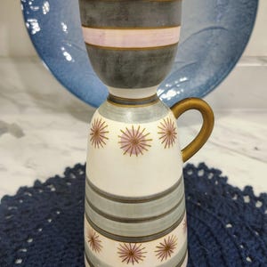 May include: A ceramic pitcher with a flared top, featuring a white base decorated with horizontal grey bands, a pale pink stripe, and small starburst patterns in pink and brown. It has a gold-toned handle and gold-toned rim accents.