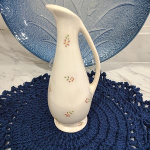 May include: A white ceramic pitcher with a curved handle and small floral accents in pink and green. The pitcher sits on a navy blue crocheted doily, with a blue decorative plate in the background.