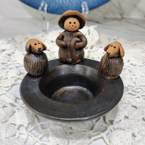 May include: A dark brown ceramic bowl featuring a central figure of a shepherd wearing a hat and holding a staff, flanked by two small sheep figures on the rim. The bowl has a smooth, dark finish and the figures have a textured, matte appearance.