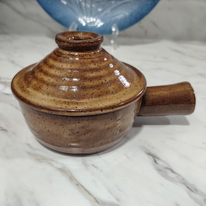 May include: A brown ceramic soup bowl with a lid and a handle. The bowl has a speckled texture and a circular lid with a small knob. The handle is attached to the side of the bowl. The bowl is on a white marble surface.