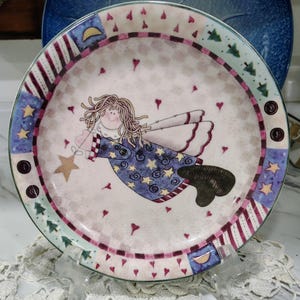 May include: A round ceramic plate featuring a central illustration of a winged figure with blonde hair wearing a blue star-patterned robe. The border displays a patchwork design with small red hearts, yellow stars, green trees, and crescent moons.