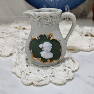 May include: A small light gray ceramic pitcher featuring a dark green oval medallion on the front. The medallion displays a white relief profile of a person and is framed by ornate gold-colored trim. The pitcher has a handle and decorative molding.