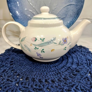 May include: A white ceramic teapot with floral designs in pink, purple, and green, set on a navy blue crocheted doily. A blue decorative plate is in the background. The teapot has a curved handle and a lid with a small knob.
