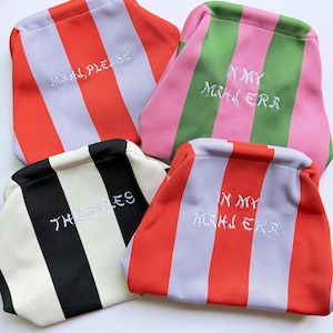 Op de afbeelding: Vier gestreepte pouches in verschillende kleurencombinaties: rood en lavendel, roze en groen, zwart en crème en rood en lavendel. Elke pouch heeft witte geborduurde tekst. De pouches zijn waarschijnlijk voor cosmetica of kleine items.