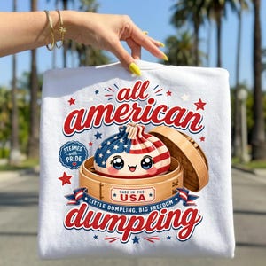 Könnte beinhalten: Weißes T-Shirt mit einem Aufdruck eines amerikanischen Flaggen-Knödels in einem Bambusdämpfer. Der Text lautet "all american dumpling" in Rot und Blau, mit den zusätzlichen Texten "Steamed with Pride" und "Made in the USA."