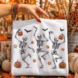 May include: A white sweatshirt featuring a graphic print of three dancing skeletons surrounded by orange autumn leaves, black bats, and jack-o'-lanterns. The skeletons are depicted in various poses, with two holding small pumpkins by their stems.
