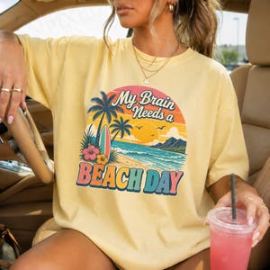 May include: A pale yellow t-shirt featuring a graphic print of a tropical beach scene with palm trees, a surfboard, and hibiscus flowers. The text reads My Brain Needs a BEACH DAY in a retro font, set against a circular sunset background.
