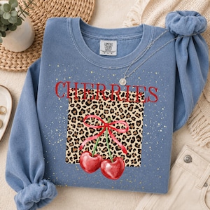 Op de afbeelding: Een blauwe sweater met ronde hals en een centraal vierkant grafisch ontwerp met een luipaardprint. Het woord CHERRIES staat in het rood boven het vierkant gedrukt. Twee rode kersen met een rode strik staan centraal op de luipaardprint.
