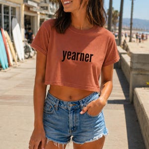 Op de afbeelding: Een terracotta-kleurige crop top met korte mouwen en het woord "yearner" in zwarte schreefloze letters op de borst, gecombineerd met een blauwe spijkerbroek met een gerafelde zoom en een klassiek ontwerp met vijf zakken.