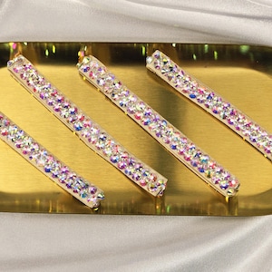 May include: Four crystal-covered hair clips are arranged on a gold-coloured oval tray. The clips are rectangular and adorned with iridescent crystals. The tray rests on a white satin surface. The logo "KRI DESIGN" is in the bottom right corner.