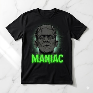 May include: Black t-shirt featuring a graphic of Frankenstein's monster with the word "MANIAC" in green, jagged lettering. The monster's face is gray with stitched scars, and the text is outlined with a green glow. The design is centered on the shirt.