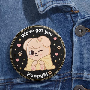 Peut inclure: Un bouton rond noir avec un dessin de chiot de dessin animé. Le chiot est enveloppé dans une couverture jaune et porte un pansement sur la tête. Le bouton porte l'inscription "We've got you" et "PuppyM" en blanc.