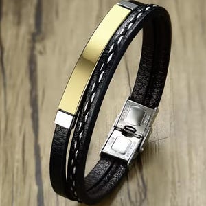 Könnte beinhalten: Ein schwarzes Lederarmband mit einer goldfarbenen rechteckigen Platte. Das Armband hat einen geflochtenen weiß-schwarzen Abschnitt und einen Edelstahlverschluss. Das Armband ist mehrschichtig gestaltet.