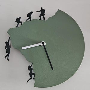 Op de afbeelding: Een ronde wandklok met een matte saliegroene afwerking en een grillige, bergachtige rand. Vijf zwarte silhouetfiguren van rotsklimmers zijn langs de onregelmatige omtrek geplaatst. De klok heeft één witte wijzer en één zwarte wijzer.