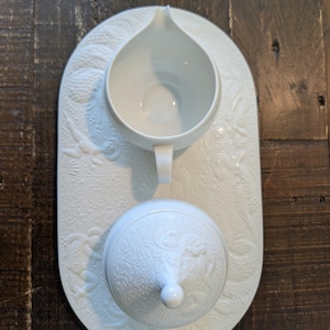May include: A white porcelain creamer and covered sugar bowl set on a matching oval tray. The set features a raised floral and botanical design. The creamer has a handle and spout. The sugar bowl has a knob on top of the lid.