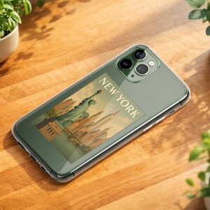 May include: A clear phone case with a New York City design featuring the Statue of Liberty and the text "NEW YORK." The phone case is on a wooden surface, surrounded by potted plants.