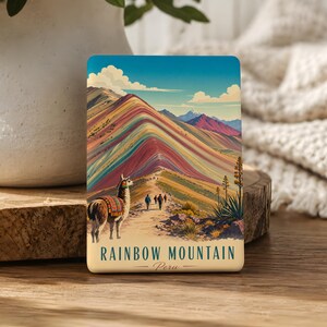 May include: A rectangular magnet featuring a colorful illustration of Rainbow Mountain in Peru. The image shows a striped mountain range, a llama in the foreground, and hikers on a path. The text reads RAINBOW MOUNTAIN Peru in dark lettering.