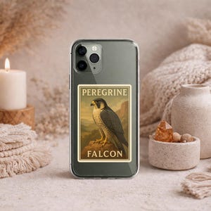 May include: A clear phone case featuring a rectangular sticker with a vintage illustration of a Peregrine Falcon. The artwork depicts the bird perched on a rock with the words "PEREGRINE FALCON" above and below the image. The case is surrounded by decorative items.