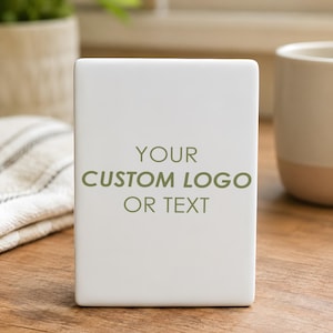 May include: A white rectangular ceramic item with the text "YOUR CUSTOM LOGO OR TEXT" in green. The item is on a wooden surface, with a white and beige mug and a plant in the background.