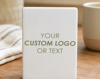 May include: A white rectangular ceramic item with the text "YOUR CUSTOM LOGO OR TEXT" in green. The item is on a wooden surface, with a white and beige mug and a plant in the background.