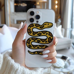 May include: Clear phone case featuring a yellow and brown snake design. The snake is coiled and has a detailed pattern. The case is held in a hand with long, manicured nails. The phone case is a protective accessory.
