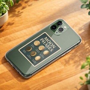 May include: A green phone case with a "Moon Phases" graphic, displaying the different phases of the moon. The phone case is on a wooden surface, with small potted plants in the background.