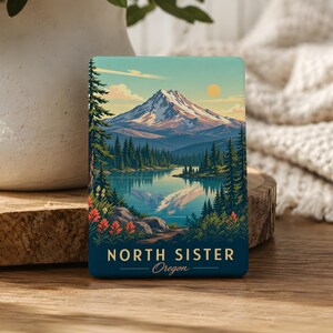 May include: A rectangular magnet featuring a colorful illustration of North Sister mountain in Oregon. The scene depicts a snow-capped peak, a calm lake reflecting the mountain, evergreen trees, and orange wildflowers. The text reads NORTH SISTER Oregon.