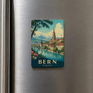 May include: A rectangular magnet featuring a scenic illustration of Bern, Switzerland. The artwork depicts a river, buildings, a church, and mountains under a blue sky. The word "BERN" and "Switzerland" are printed on the magnet.
