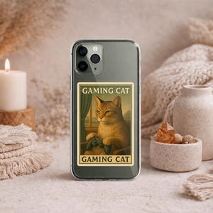 May include: Clear phone case featuring a framed image of a cat holding a game controller, with the text "GAMING CAT" above and below the image. The cat is orange and white, and the case is surrounded by neutral-toned decor.