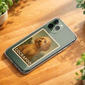May include: A clear phone case with a Cockapoo design. The case features a rectangular image of a Cockapoo dog with the word "COCKAPOO" in a white font. The phone case is on a wooden surface with small potted plants.