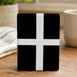 May include: A black rectangular object with a white cross design. The object is on a wooden surface, with a white mug and a plant in the background. The cross is centered on the object.