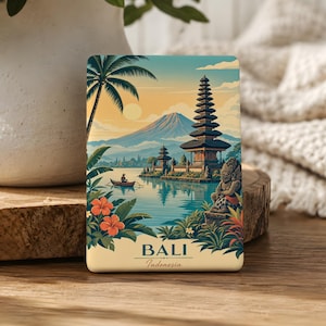 May include: A rectangular magnet featuring a scenic illustration of Bali, Indonesia. The artwork depicts a traditional Balinese temple, a mountain, and a boat on the water, with palm trees and orange flowers. The word "BALI" is printed at the bottom.
