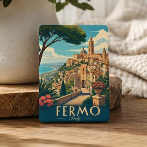 May include: A rectangular metal sign featuring a colorful illustration of the Italian city of Fermo, with a coastal landscape, historic stone buildings, a tall bell tower, and pink flowers. The text FERMO Italy is printed in white at the bottom.