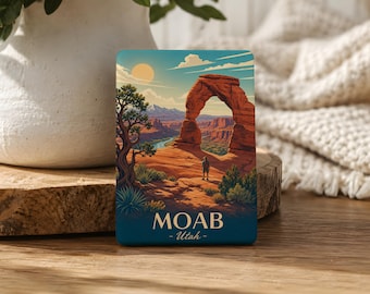 May include: A rectangular magnet featuring a scenic illustration of Delicate Arch in Moab, Utah. The artwork depicts a person standing before the arch, with a river and mountains in the background. The text "MOAB - Utah" is at the bottom.