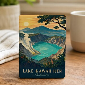 May include: A rectangular magnet featuring a vibrant illustration of Lake Kawah Ijen in Indonesia. The artwork depicts a turquoise crater lake surrounded by rugged mountains, steam, and foliage under a sunset sky. The text reads Lake Kawah Ijen Indonesia.