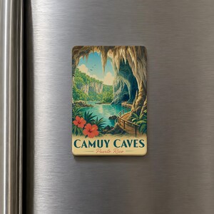 May include: A rectangular refrigerator magnet featuring a colorful illustration of the Camuy Caves in Puerto Rico. The design depicts a cavern entrance with stalactites, a blue river, lush green foliage, red hibiscus flowers, and a wooden walkway.